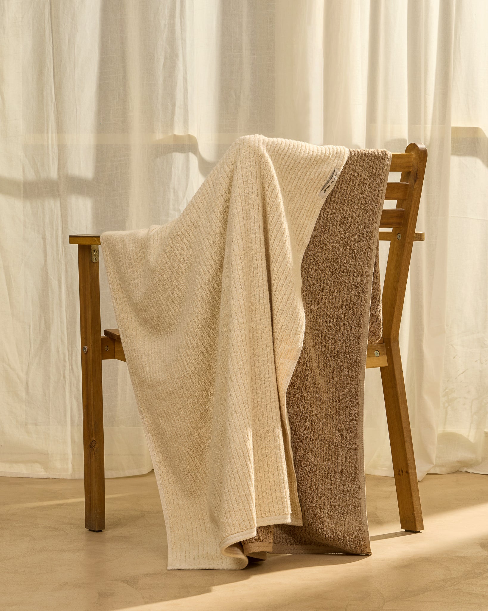 Two textured knit blankets, one cream and one taupe, draped over a wooden chair in front of sheer white curtains.